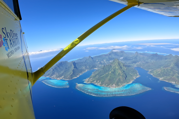 View of mountain island and turquoise waters from inside a yellow plane, labeled 'Skydive Tahiti'
