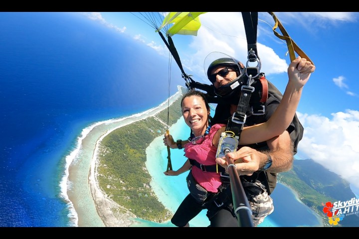 Smiling woman tandem skydiving with an instructor over a turquoise ocean and tropical island.