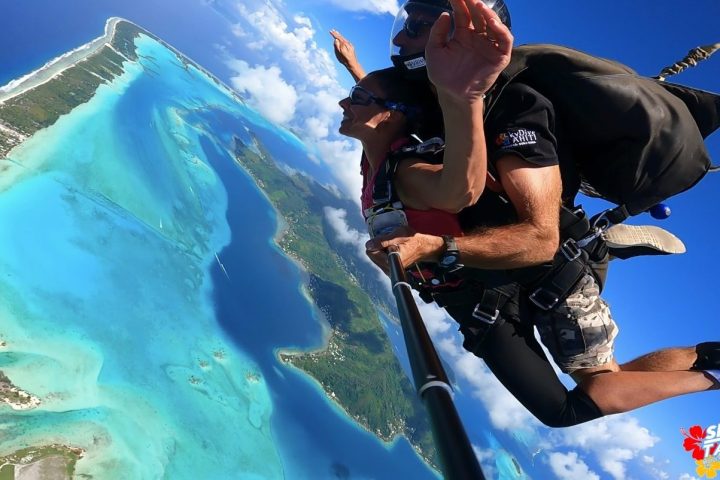 Two people tandem skydiving over a tropical island with clear blue water below.
