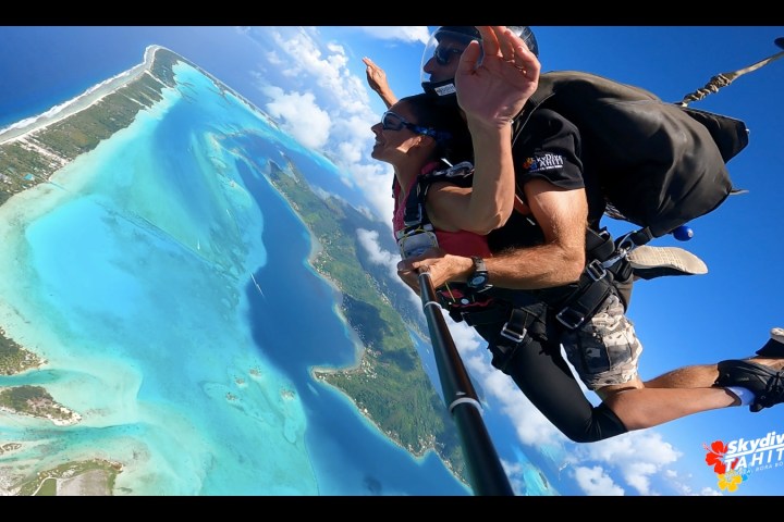 Two people tandem skydiving over a tropical island with clear blue water below.