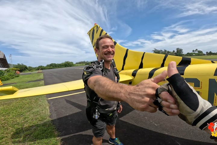 Smiling man in skydiving gear gives a thumbs-up near a yellow plane with black stripes.