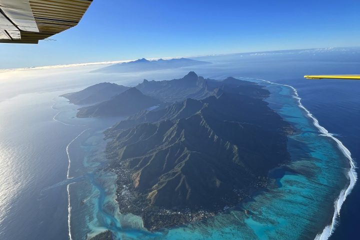 Aerial view of an island with mountains surrounded by blue ocean and reef, partial airplane wing visible.