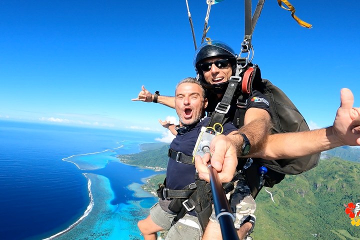 Two people tandem skydiving with ocean and coastline below, holding a selfie stick.