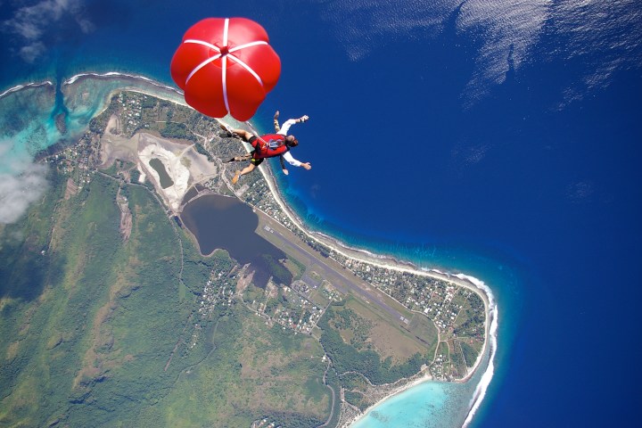 Skydiver with red parachute over coastal landscape with ocean and greenery.