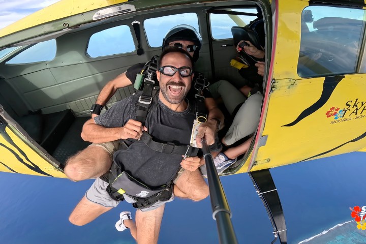 Skydivers exiting a yellow plane with 'Skydive Tahiti' logo, man smiling with selfie stick over ocean.