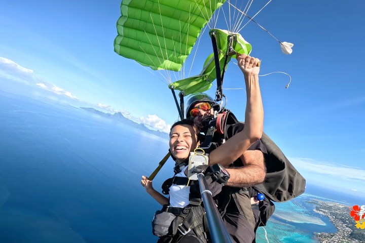 Tandem skydiving over ocean, green parachute, person smiling and holding a selfie stick, clear blue sky.